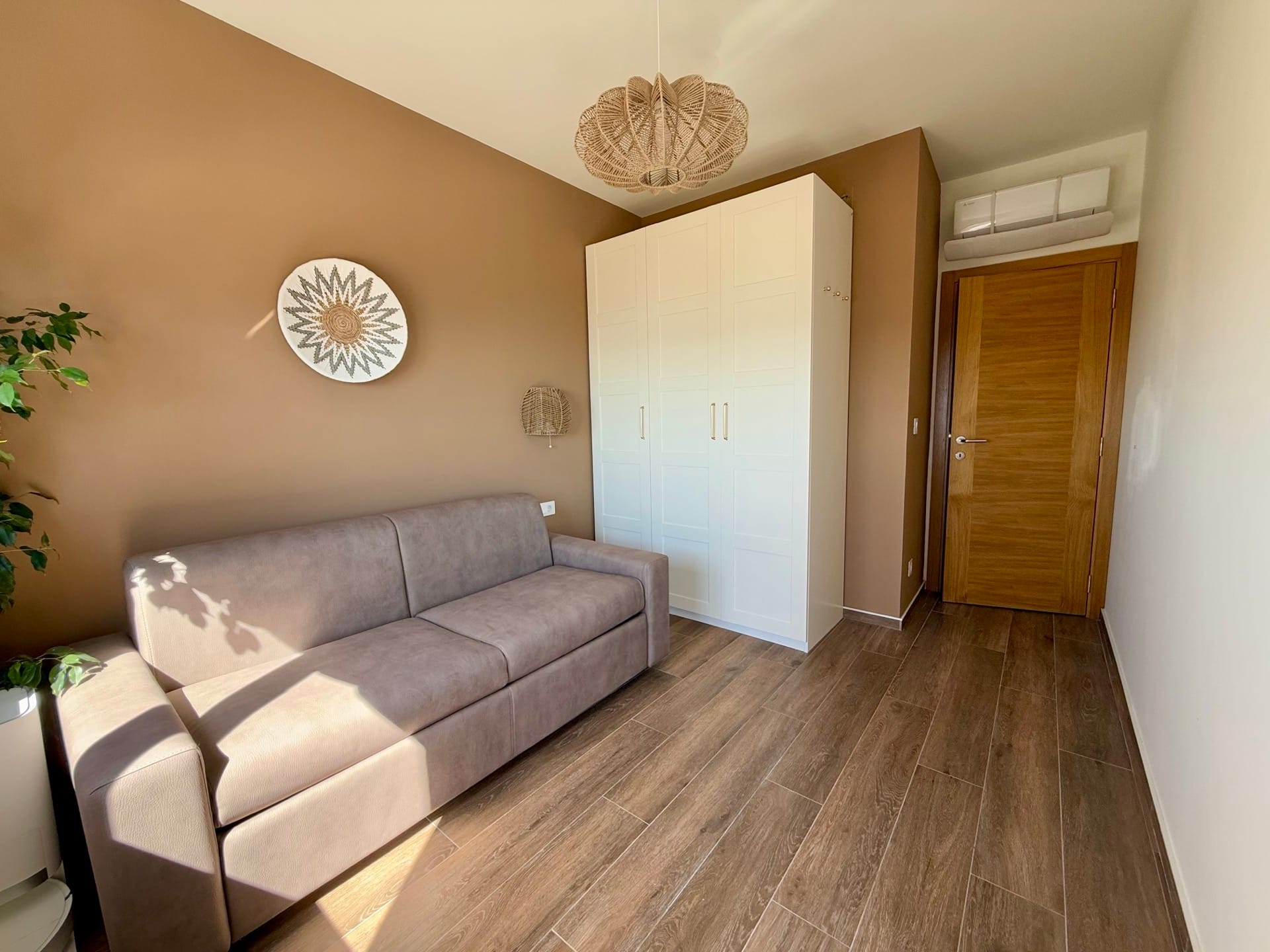 Dnevna soba u stanu u Bečićima, Crna Gora: bež sofa, beli ormar, drveni pod i pletena lampa.