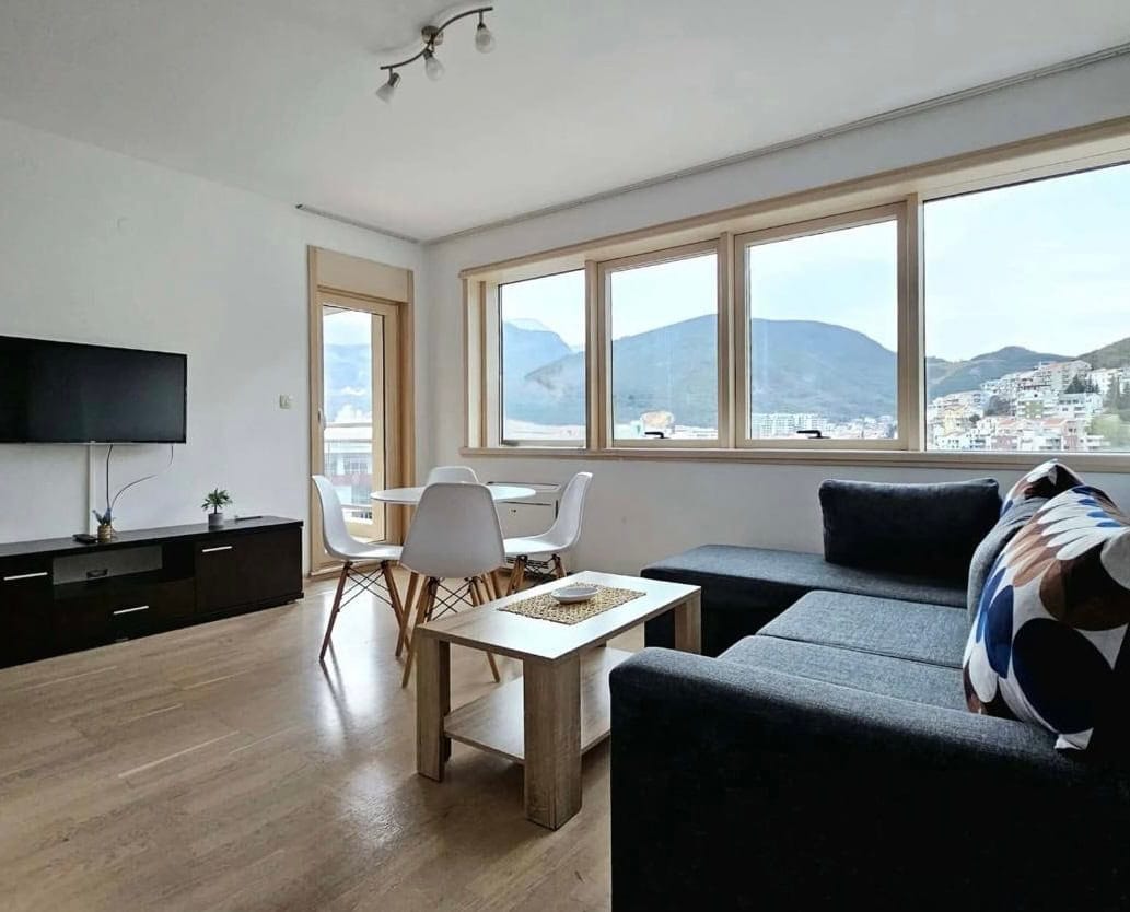 Svetao dnevni boravak u stanu u Bečićima, Crna Gora: siva sofa, trpezarijski deo i panoramski pogled na planine kroz prozore.