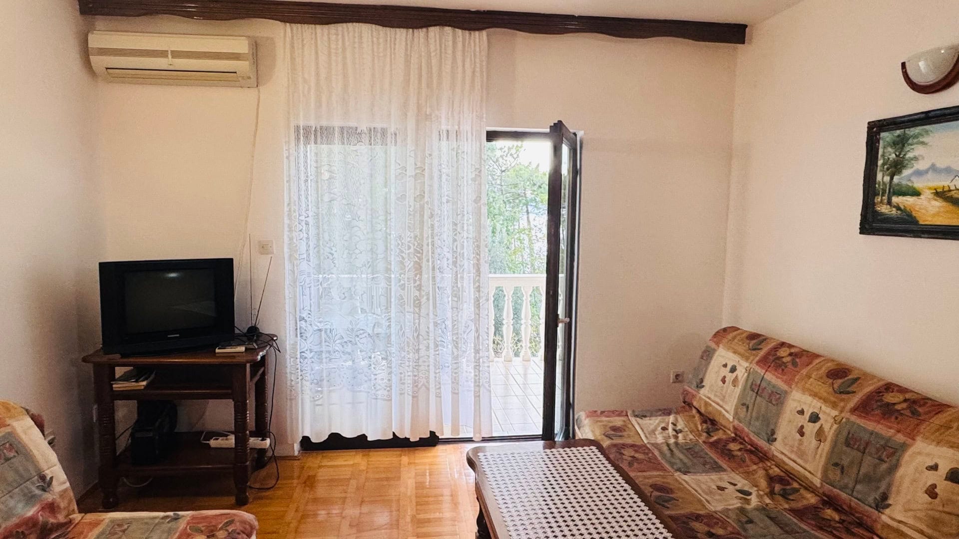 Dnevna soba u stanu na Svetom Stefanu, Crna Gora: sofa, TV, parket i izlaz na balkon s pogledom na zelenilo.