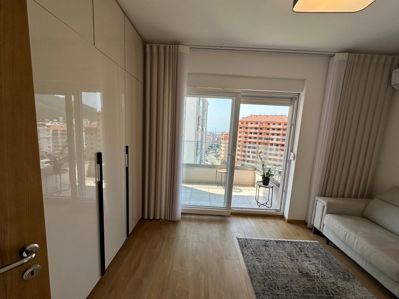 Savremeni dnevni boravak u stanu u Budvi, Crna Gora: ugradni sjajni ormar, svetla sofa i izlaz na balkon.