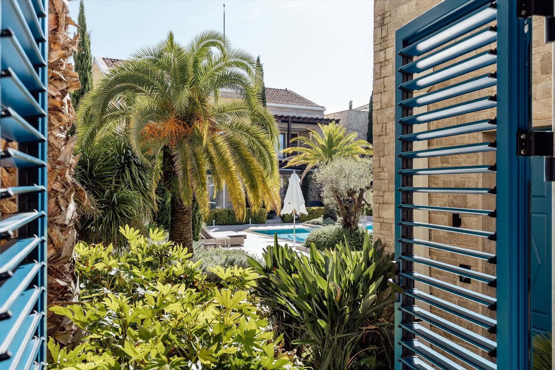 Вид из окна квартиры в Портонови, Кумбор, Черногория: бассейн, пальмы и терраса с синими жалюзи в средиземноморском стиле