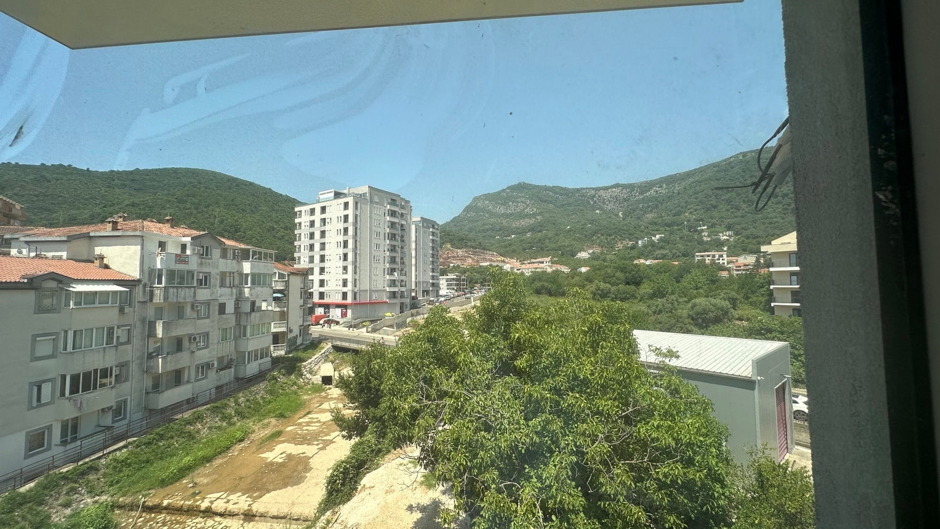 Pogled sa balkona stana u Budvi, Crna Gora: panorama grada, zelene planine i stambene zgrade po sunčanom danu.