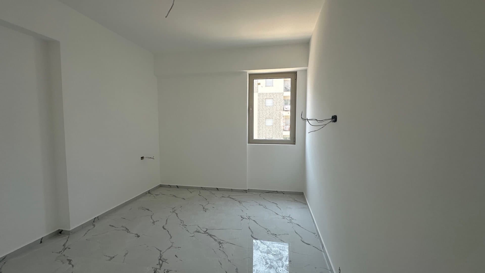 Svetla spavaća soba u novogradnji u Budvi, Crna Gora: beli zidovi, pod od porcelanskog pločica sa mermernim dezenom, pogled kroz prozor.