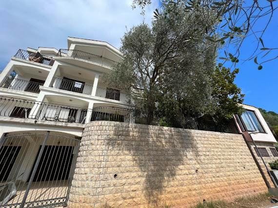 Trospratna vila u Budvi, Crna Gora: fasada sa balkonima, kameni potporni zid i maslina kod ulaza.