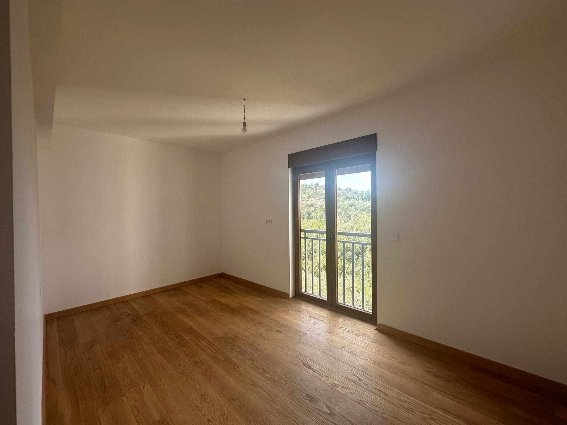 Svetla spavaća soba u stanu, Krašići, Crna Gora: parketni pod, beli zidovi i izlaz na balkon s pogledom na šumu.