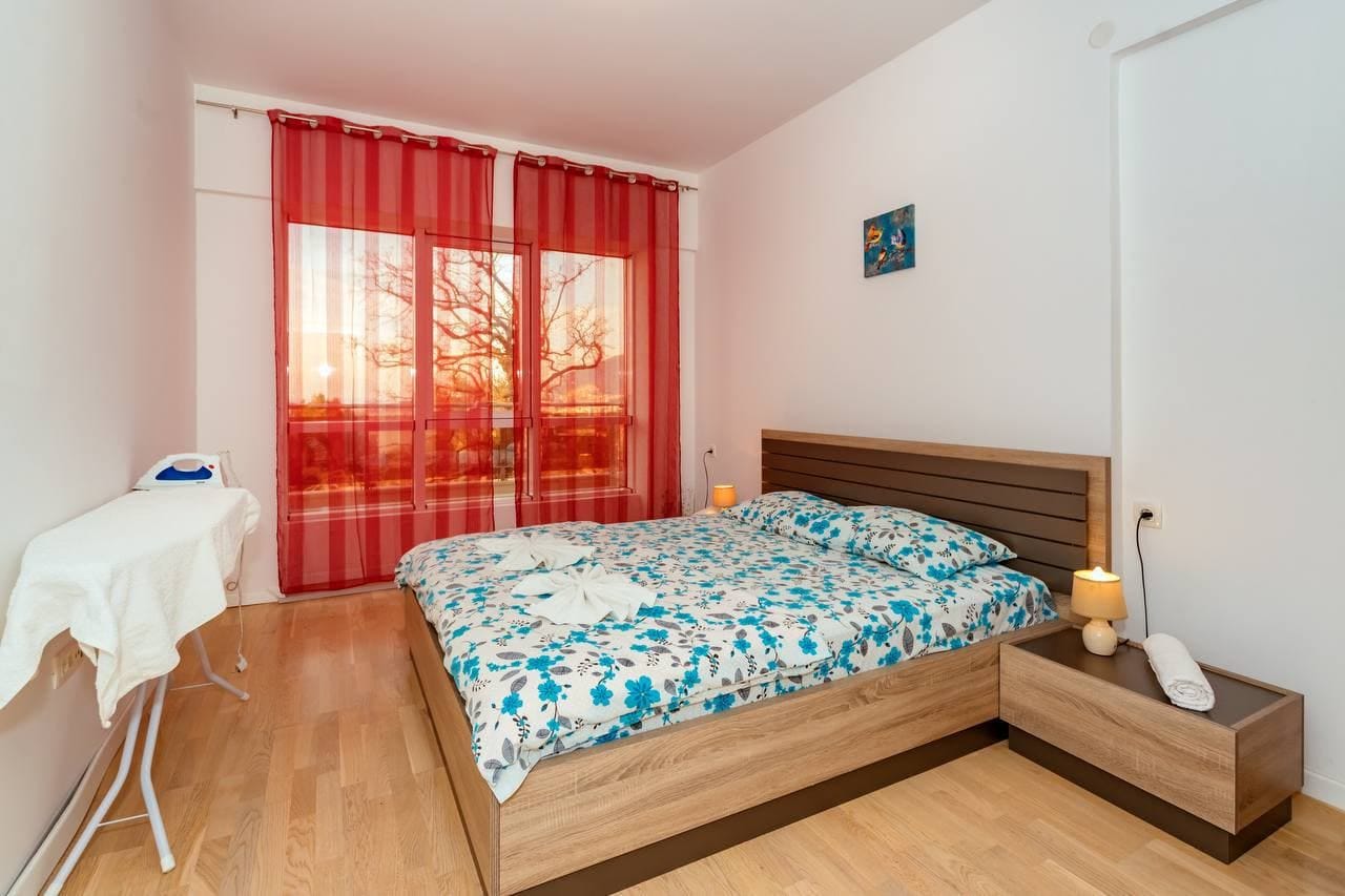 Udobna spavaća soba sa bračnim krevetom u stanu u Bečićima, Crna Gora: svetao enterijer, parket i crvene zavese.