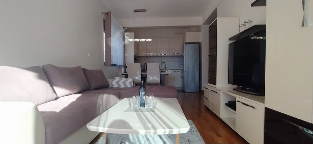 Moderna dnevna soba sa kuhinjom u stanu u Bečićima, Crna Gora: ugaona sofa, trpezarijski prostor i svetli nameštaj.