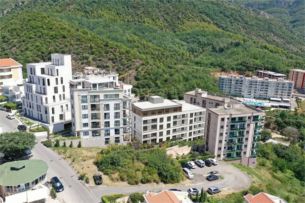 Pogled iz vazduha na moderne stambene komplekse u Bečićima, Crna Gora, sa parkingom i zelenim planinskim padinama oko njih.