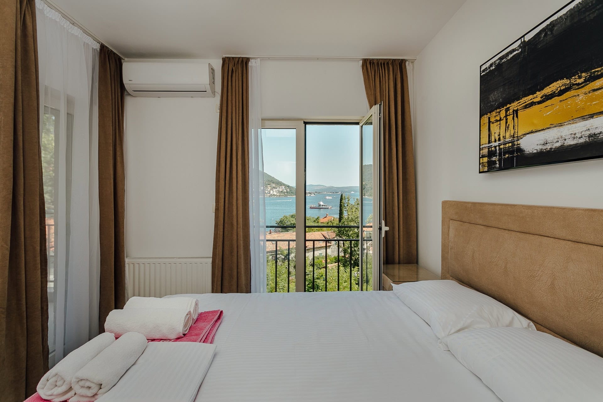 Schlafzimmer in einer Wohnung in Kamenari, Montenegro: Doppelbett, Meer- und Bergblick durch ein Panoramafenster mit Balkon.