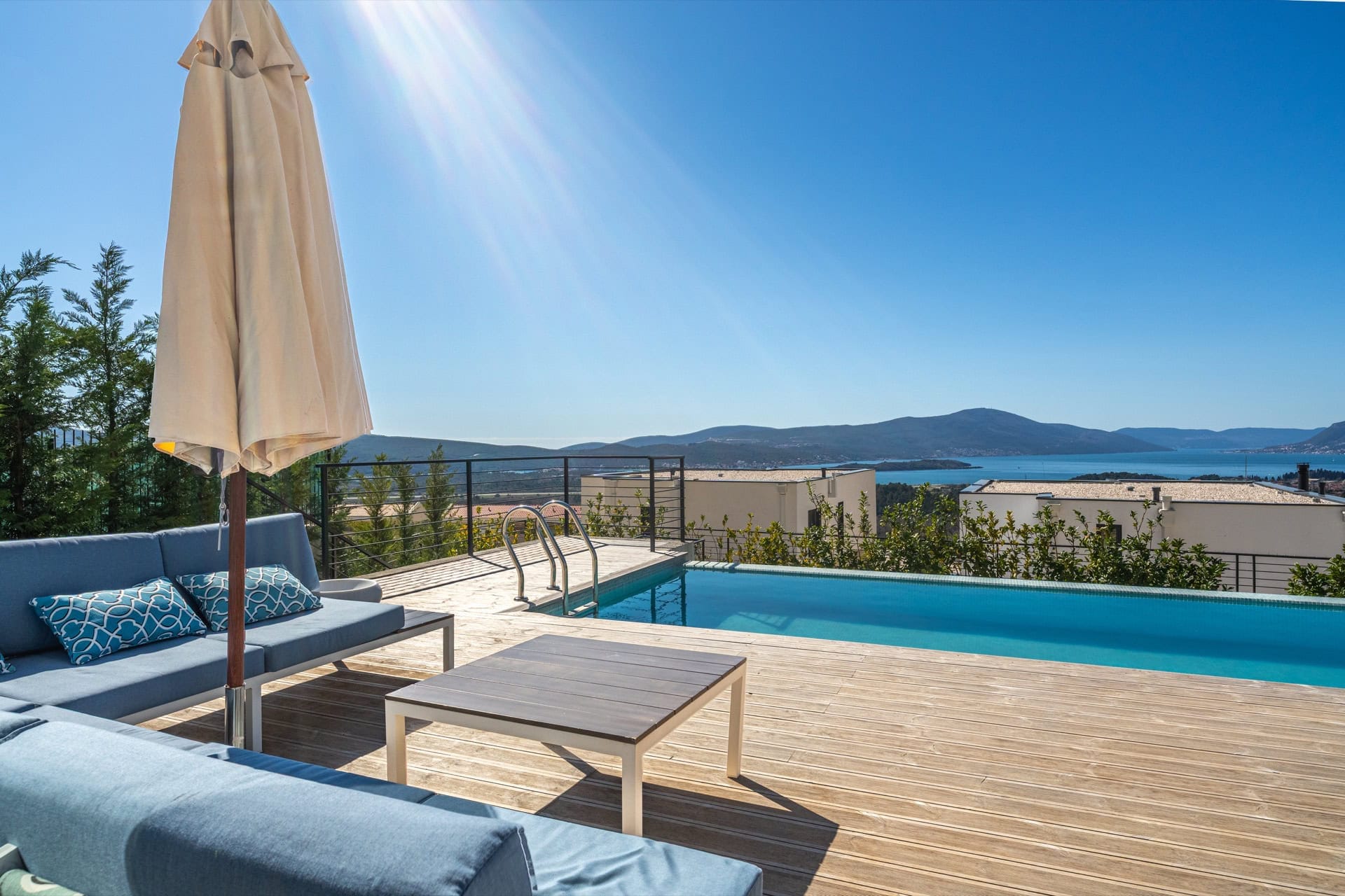 Terrace with a pool and sea view at a villa in Kavac, Montenegro: wooden deck, blue sofas, and a sun umbrella.