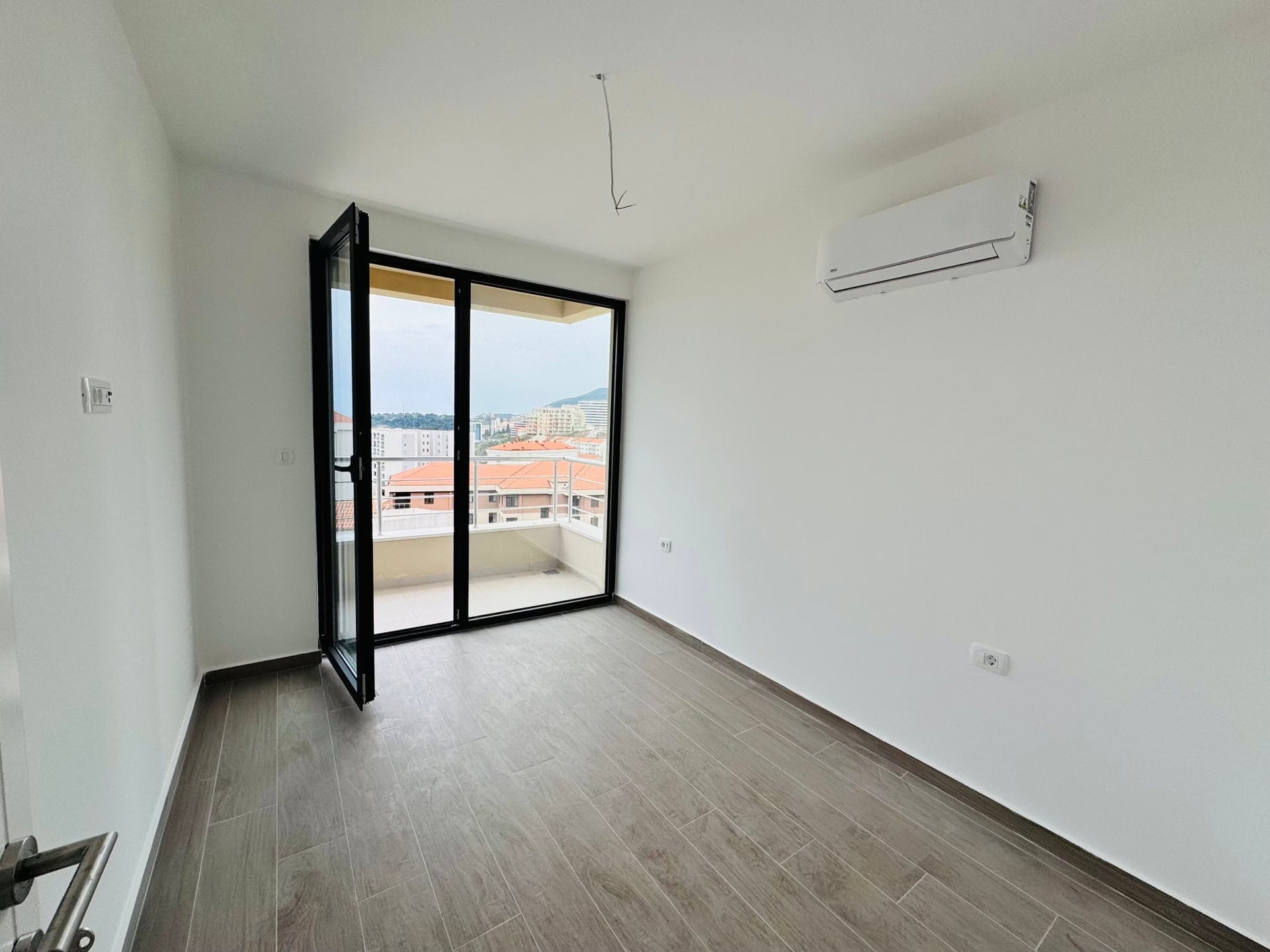 Svetla spavaća soba u stanu u Bečićima, Crna Gora: panoramski prozor, izlaz na balkon, klima-uređaj i pločice u imitaciji drveta.