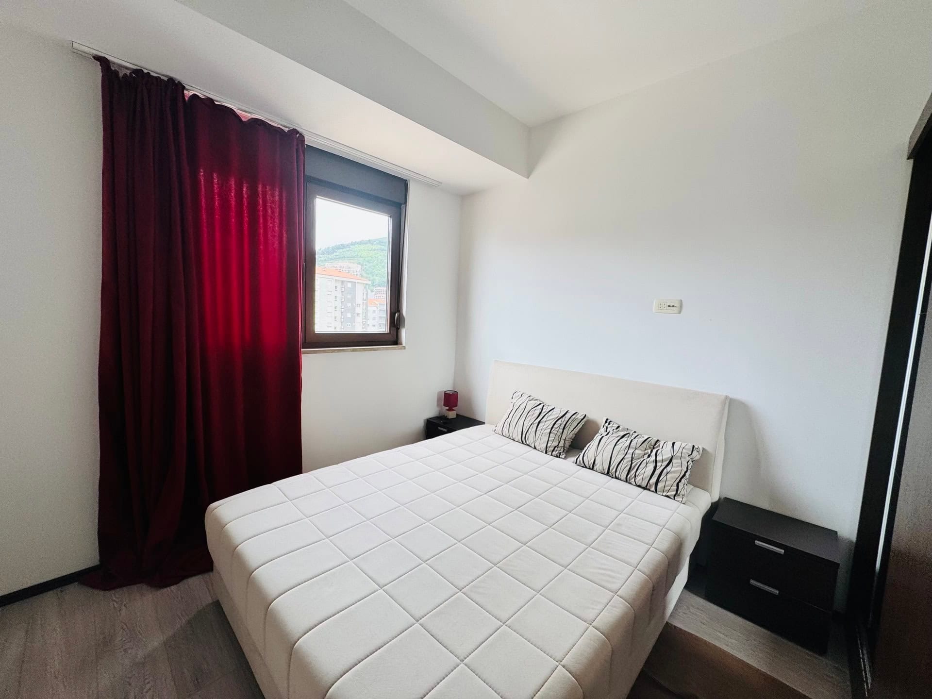 Bright bedroom with a double bed and red curtains in an apartment in Budva, Montenegro. City view from the window.