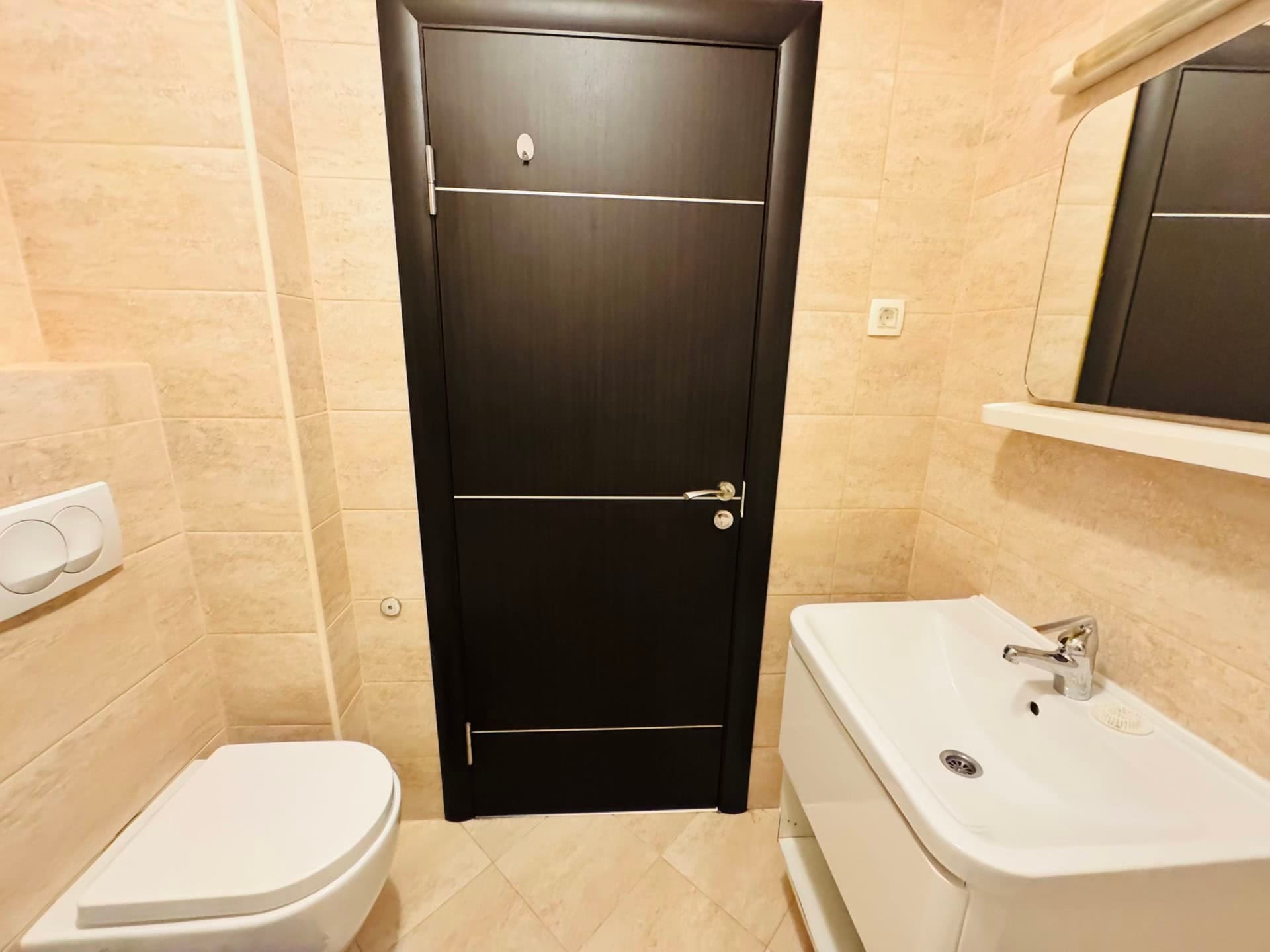 Modern bathroom in an apartment in Budva, Montenegro: white fixtures, beige tiles, and a dark interior door.