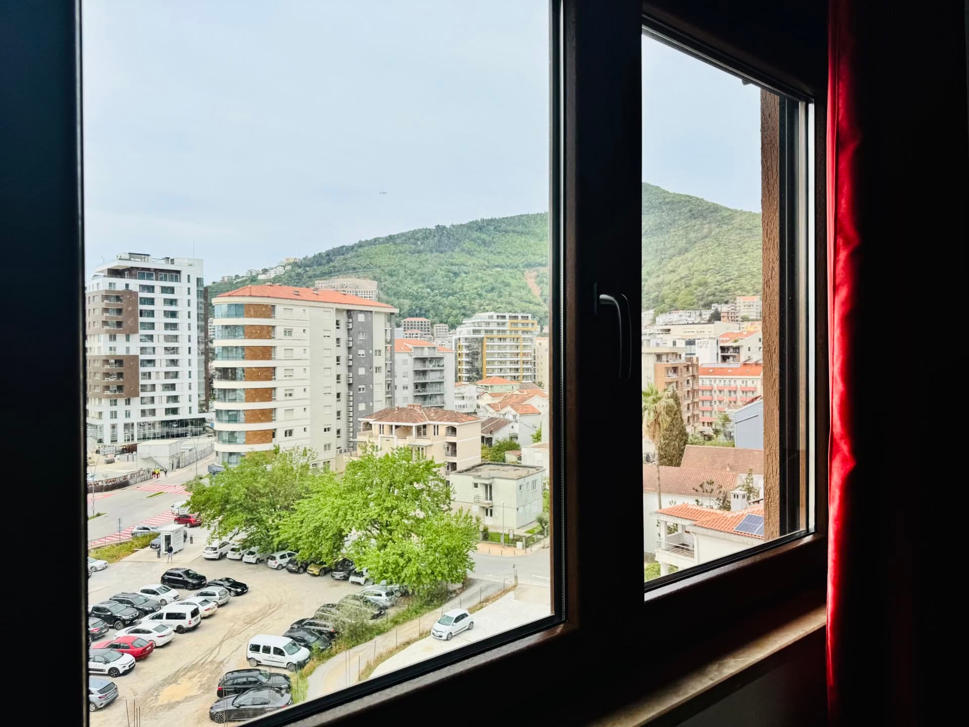 View from an apartment window in Budva, Montenegro: city panorama, residential buildings, parking lot, and green mountains in the background.