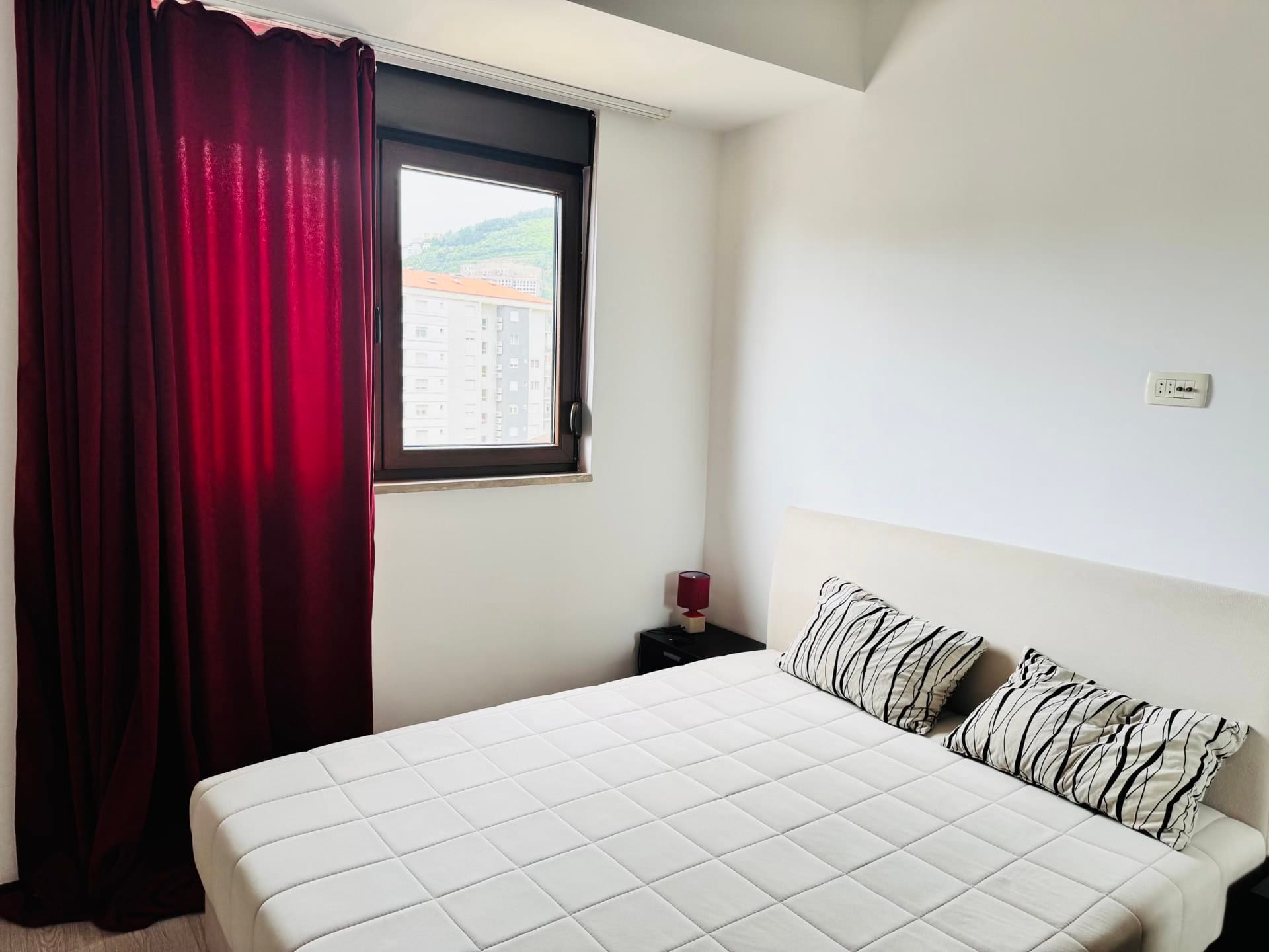 Bright bedroom with a double bed and red curtains in an apartment in Budva, Montenegro, with a city view from the window.