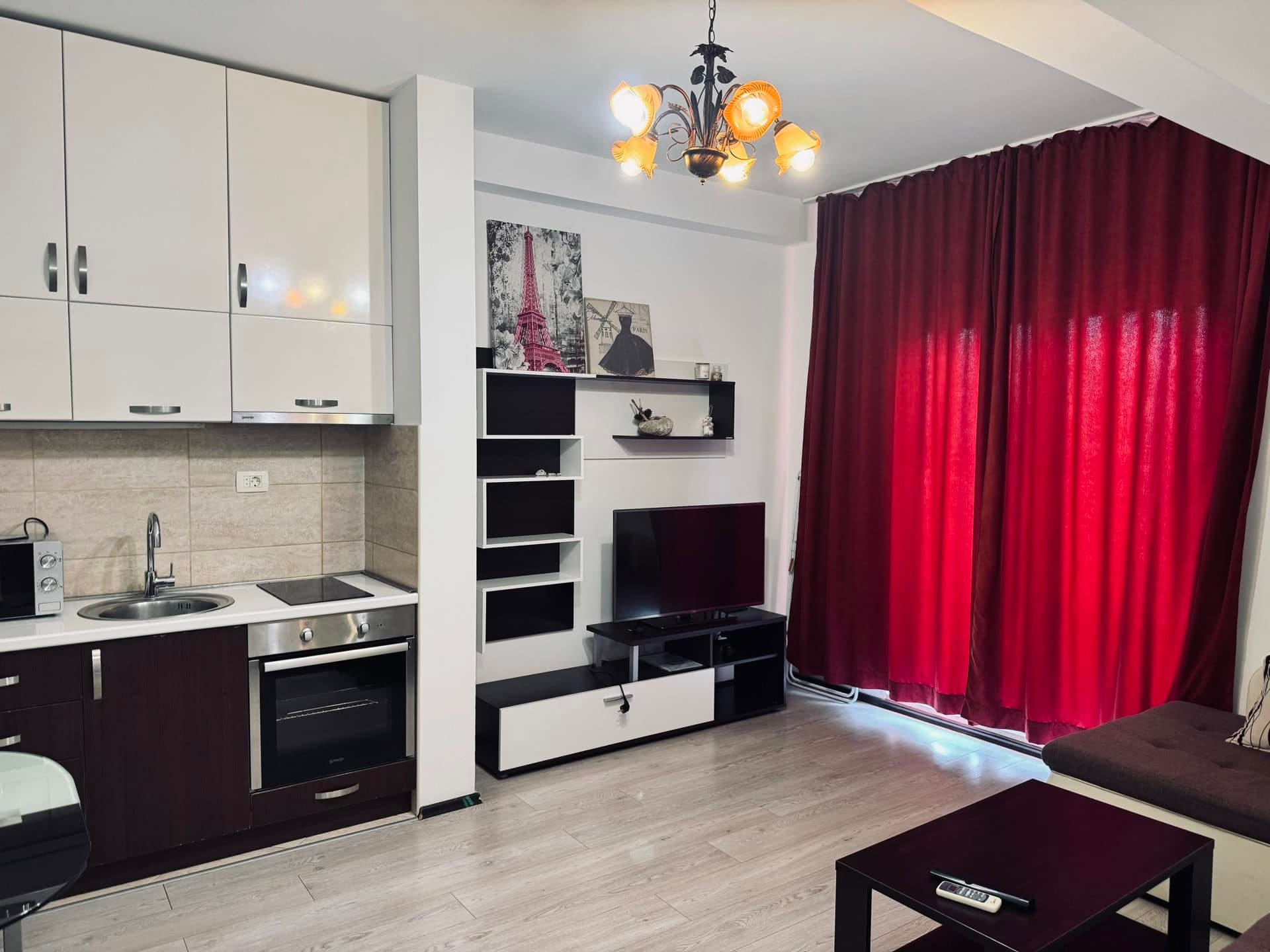 Modern living room with kitchen in an apartment in Budva, Montenegro: light furniture, TV area, and panoramic red curtains.