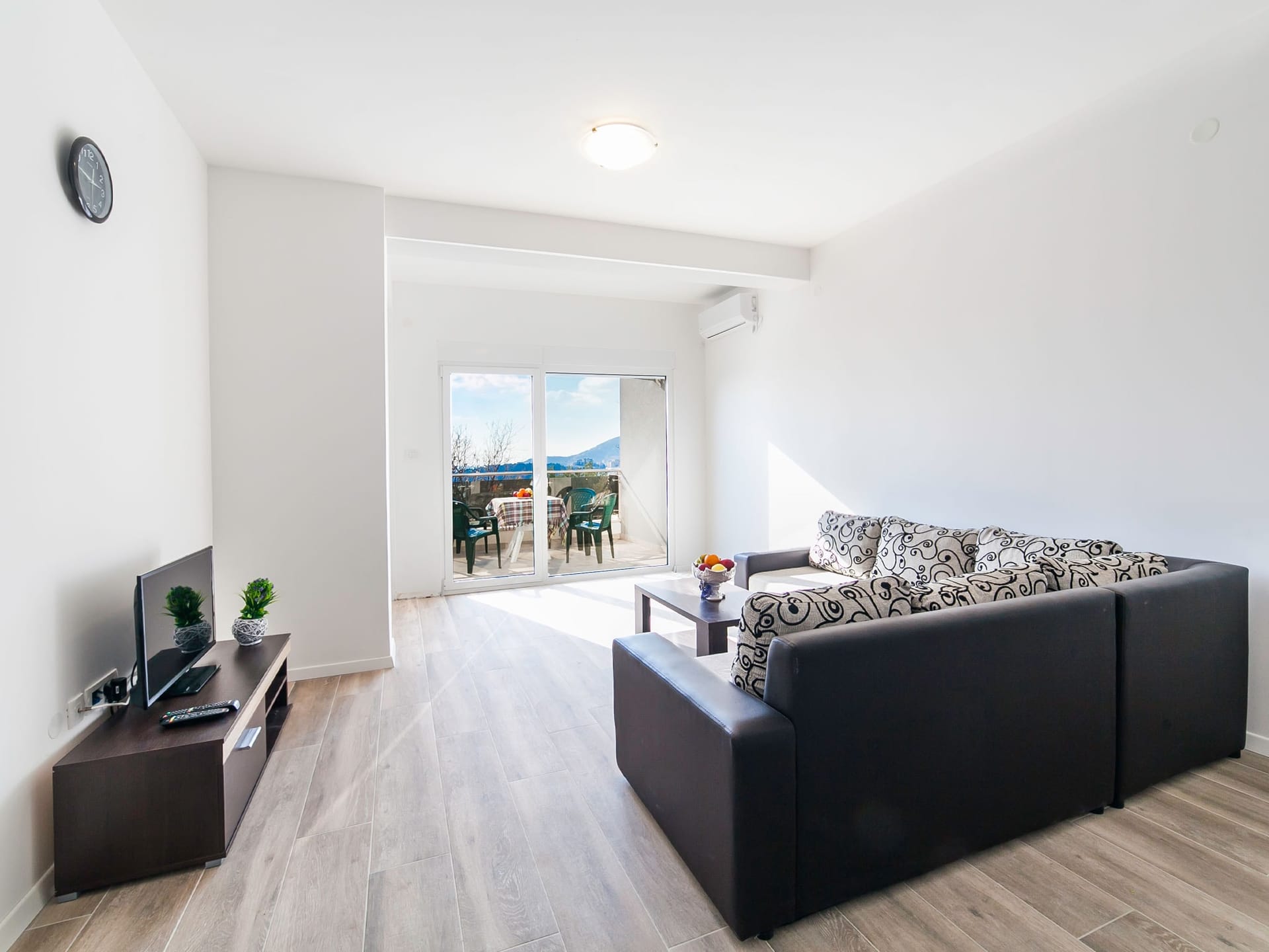 Bright living room in an apartment in Bečići, Montenegro: corner sofa, TV area, and access to a terrace with mountain views.