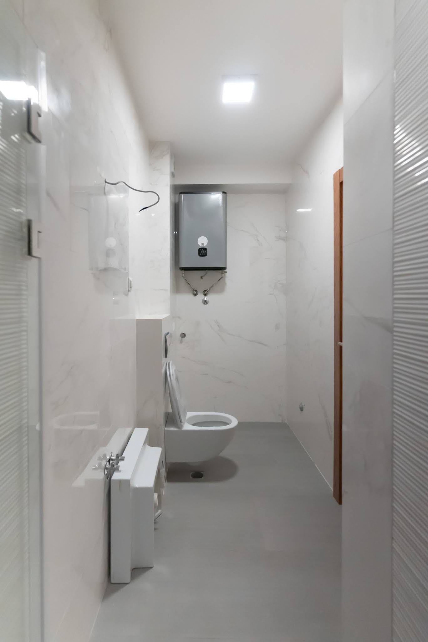Budva, Karadağ’daki bir dairede modern banyo: mermer görünümlü açık renk fayans, duvara asılı tuvalet ve бойлер.