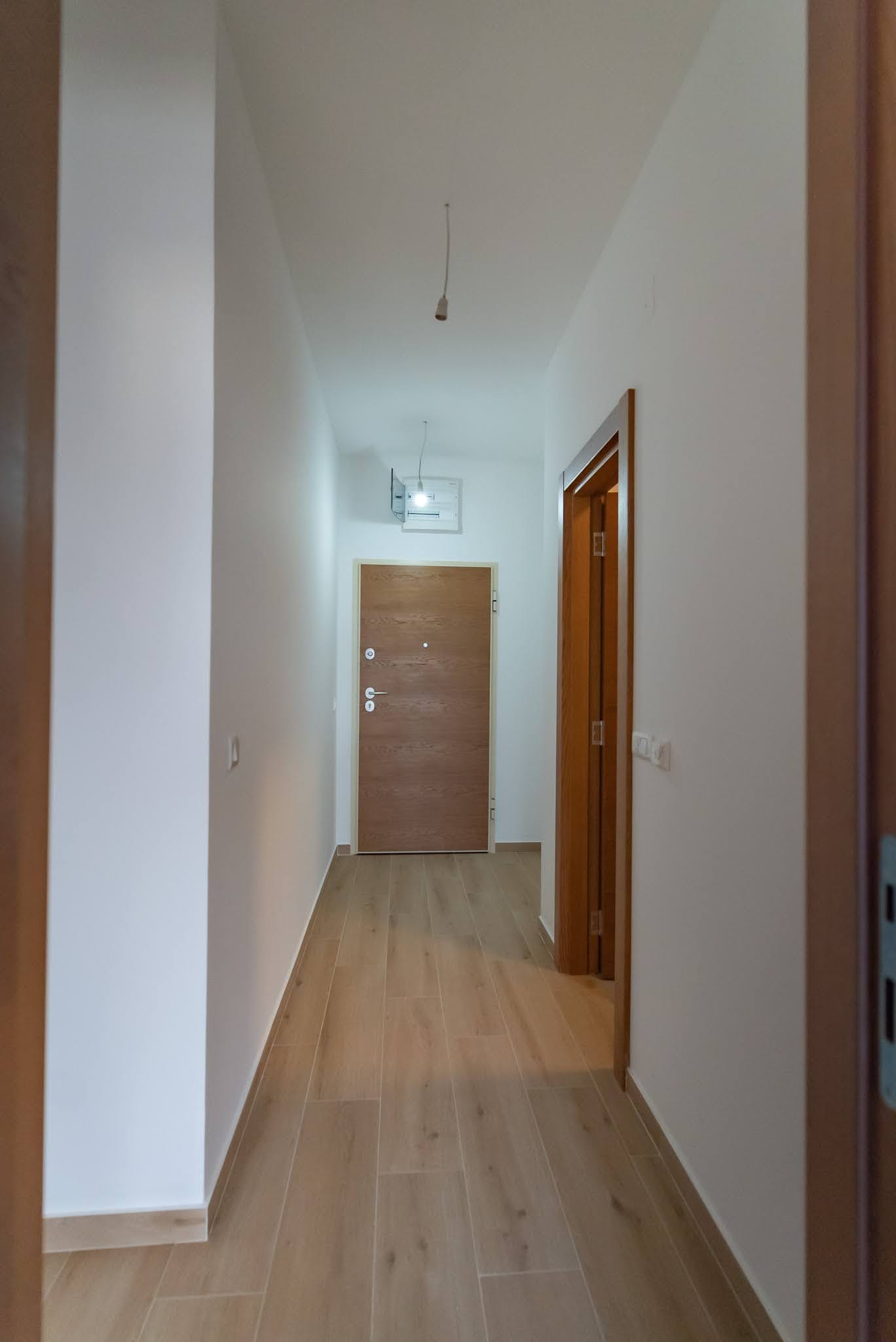 Karadağ, Budva’daki dairede aydınlık koridor: ahşap giriş kapısı, ahşap görünümlü laminat ve beyaz duvarlar.