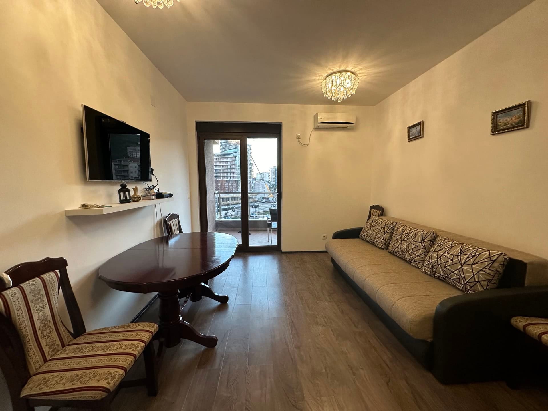Dnevna soba u stanu u Budvi, Crna Gora: sofa, trpezarijski sto, TV i izlaz na balkon s pogledom na grad.