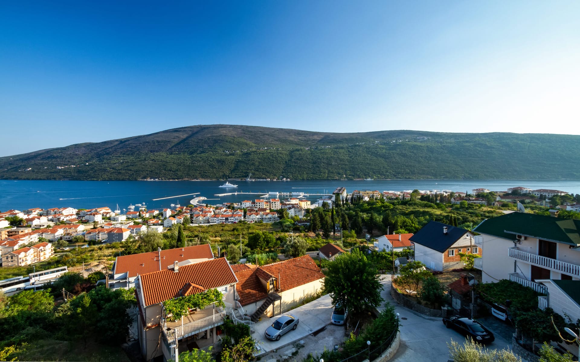 Panoramski pogled na more, zaliv i stambeni kompleks u Kumboru, Crna Gora: kuće sa crepnim krovovima i planine u pozadini.