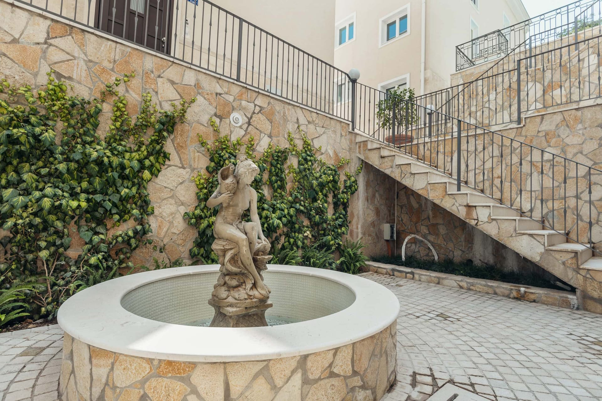 Dvorište stambenog kompleksa u Kumboru, Crna Gora: kamena obloga zidova, fontana sa statuom i stepenište do apartmana.