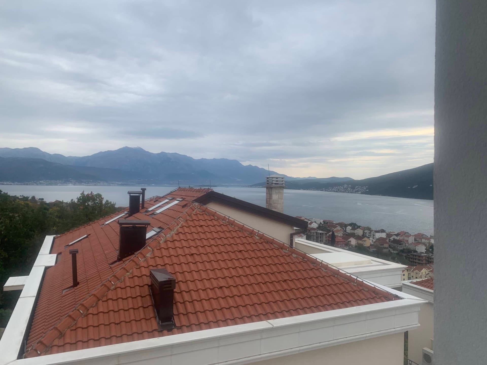 Pogled sa balkona stambenog kompleksa u Kumboru, Crna Gora: panorama mora, planina i krovova kuća sa crepovima.