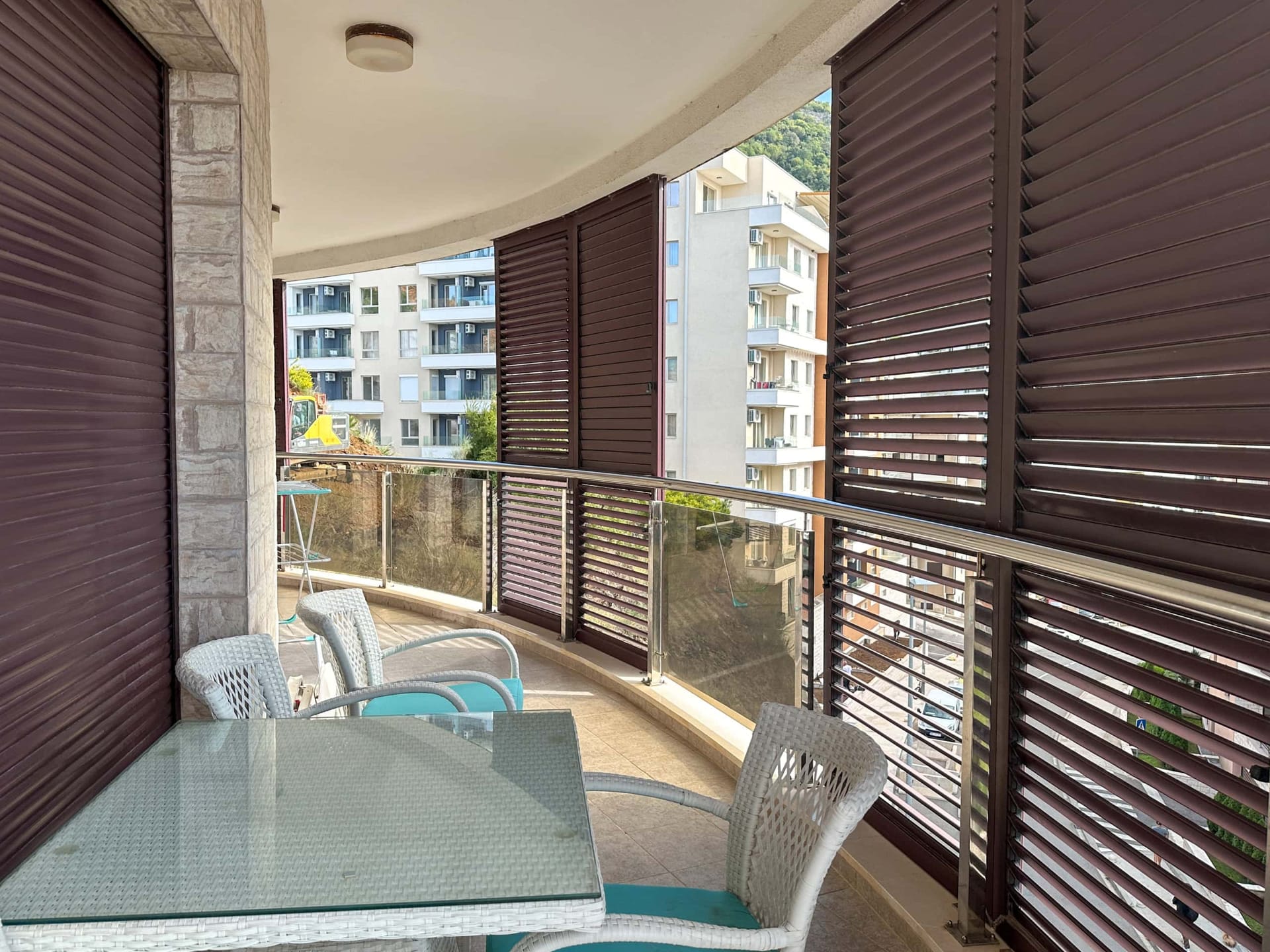 Prostran balkon sa pletenim nameštajem i zaštitnim žaluzinama u stanu u Bečićima, Crna Gora, pogled na stambeni kompleks.