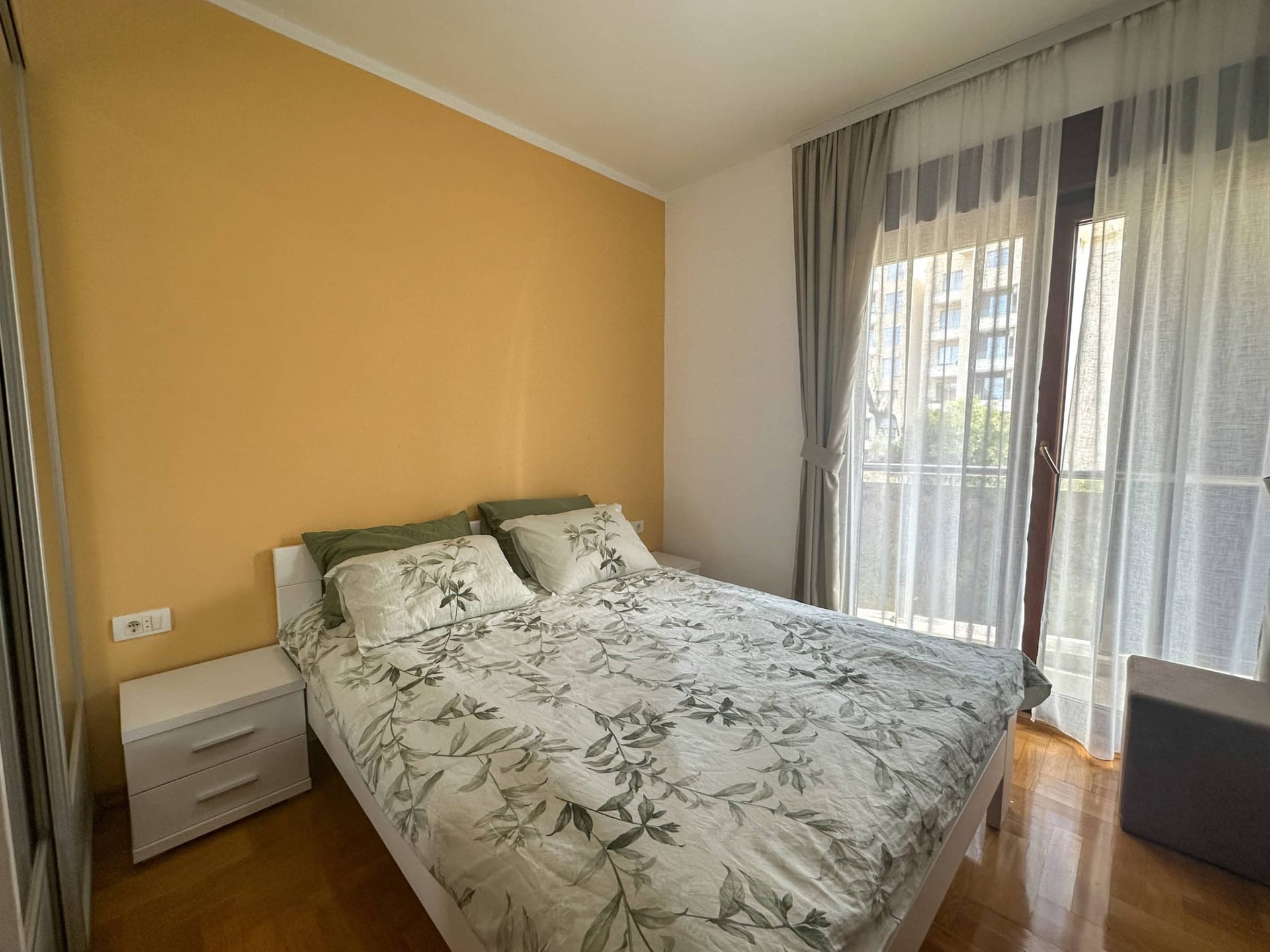 Spavaća soba sa bračnim krevetom i žutim zidom u stanu u Bečićima, Crna Gora, sa izlazom na balkon i parketom.