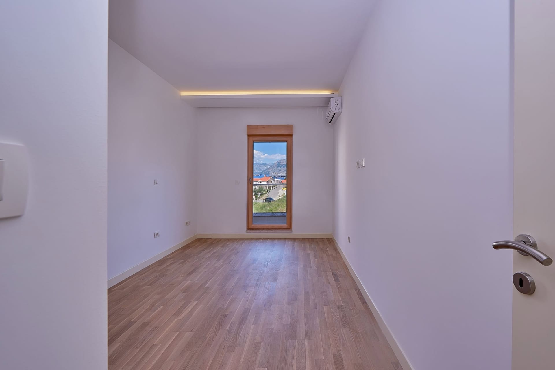 Svetla spavaća soba u stanu, Dobrota, Crna Gora: laminat, beli zidovi, klima-uređaj i pogled na planine kroz prozor.