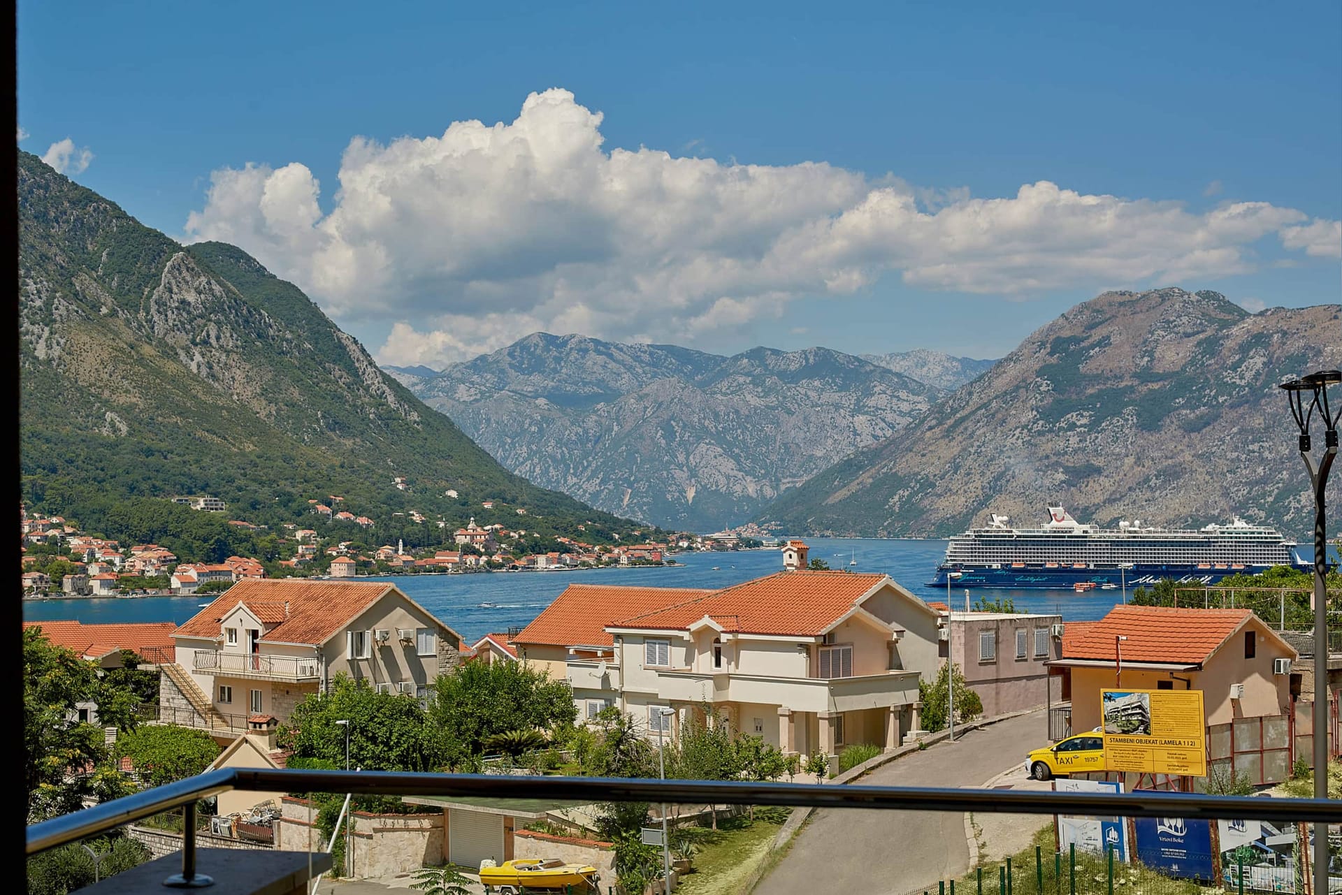 Pogled sa balkona stana u Dobroti, Crna Gora: panorama Bokokotorskog zaliva, planine, kruzer i krovovi od crepova.