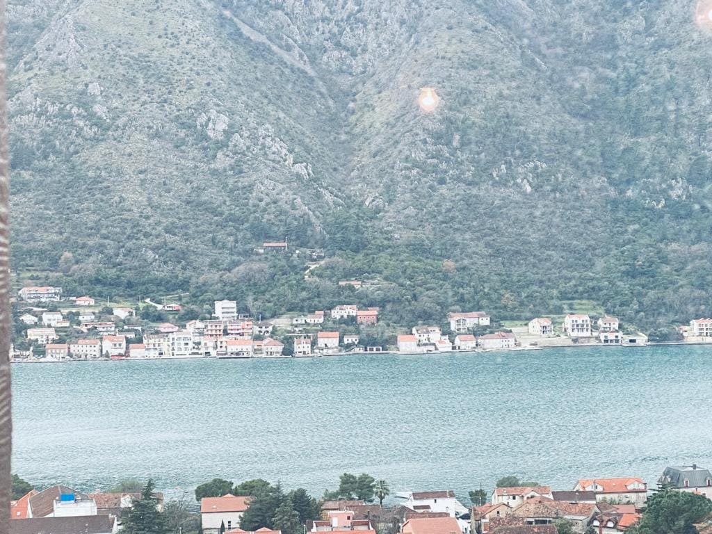Panoramski pogled na Bokokotorski zaliv i planine iz prozora stana u Dobroti, Crna Gora: primorske kuće sa crvenim krovovima.