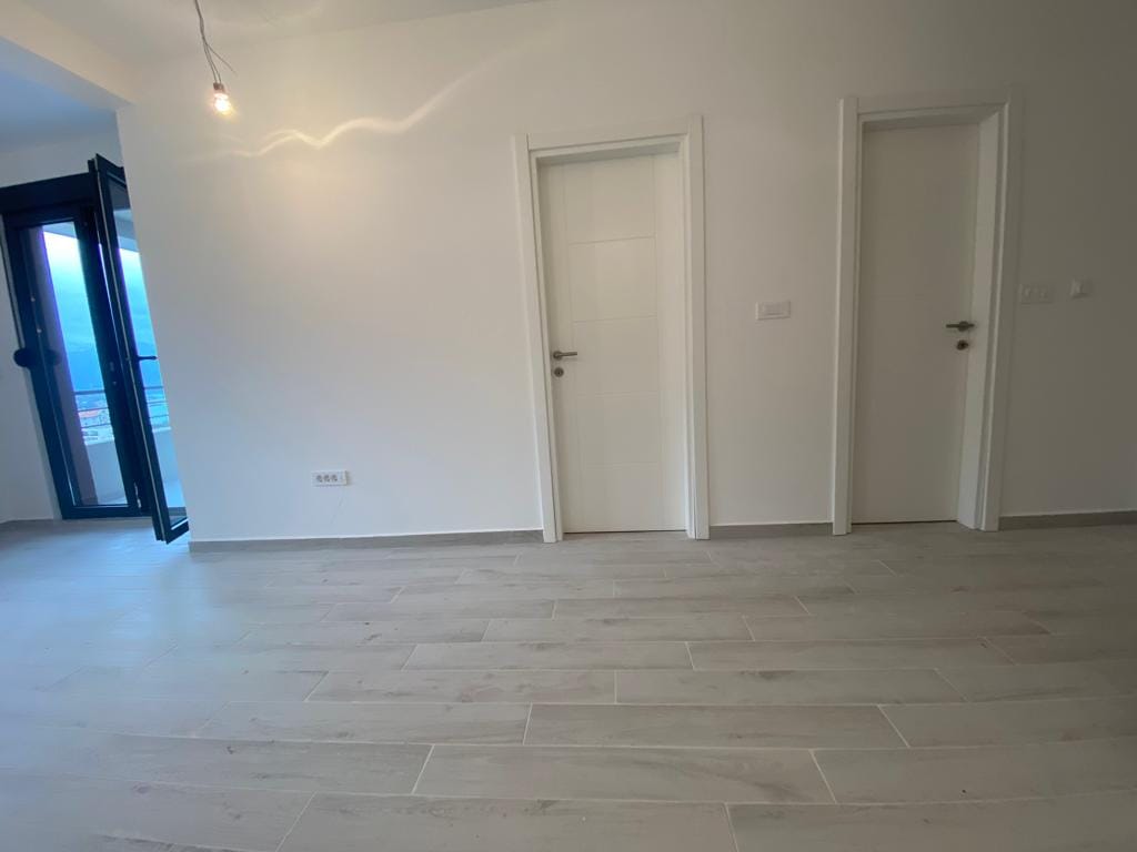 Svetla dnevna soba sa belim zidovima i podnim pločicama u novom stanu u Dobroti, Crna Gora, pogled na terasu.