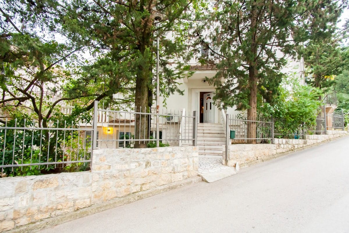 Fasada kuće u Budvi, Crna Gora: kamena ograda, metalna ograda, stepenice do ulaza i zelena stabla u dvorištu.