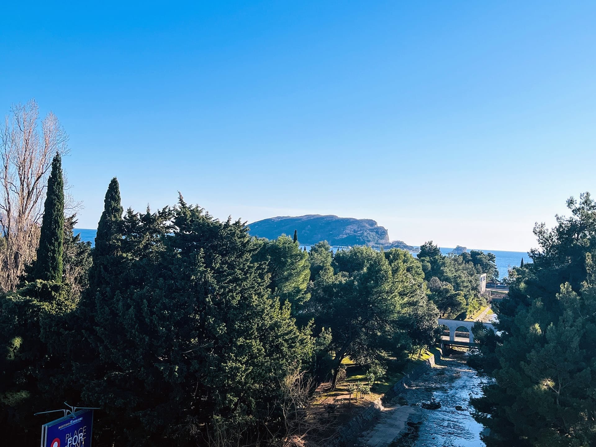 Budva, Karadağ’da deniz, dağlar ve nehirin panoramik manzarası; ön planda yeşil ağaçlar ve yaya köprüsü.