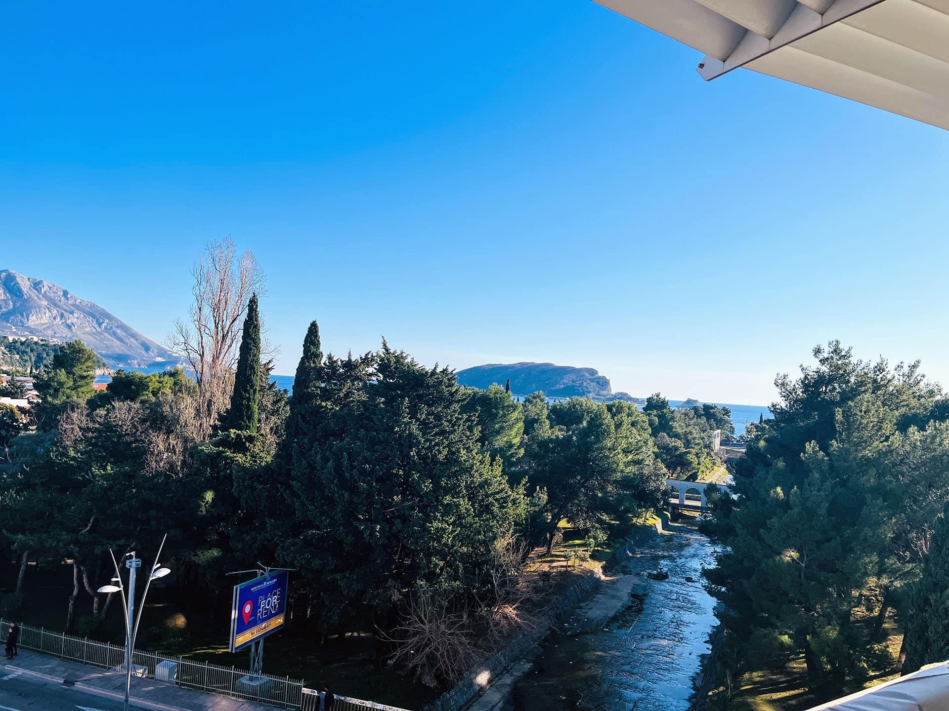 Budva, Karadağ’daki daire balkonundan görünüm: dağlar, yeşil park, nehir ve deniz kıyısının panoraması.