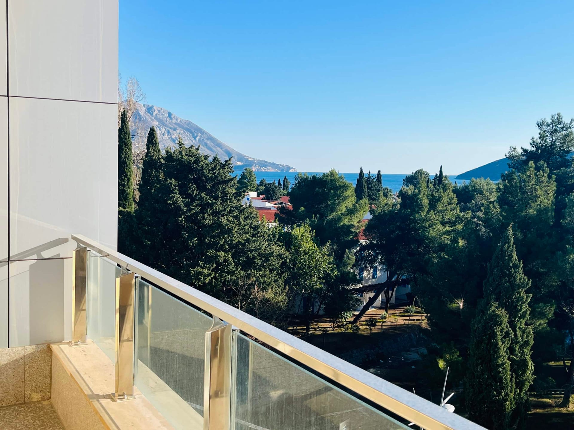 Karadağ, Budva’daki dairenin balkonundan görünüm: deniz, dağlar ve iğne yapraklı ağaçlarla yeşil bir park panoraması.