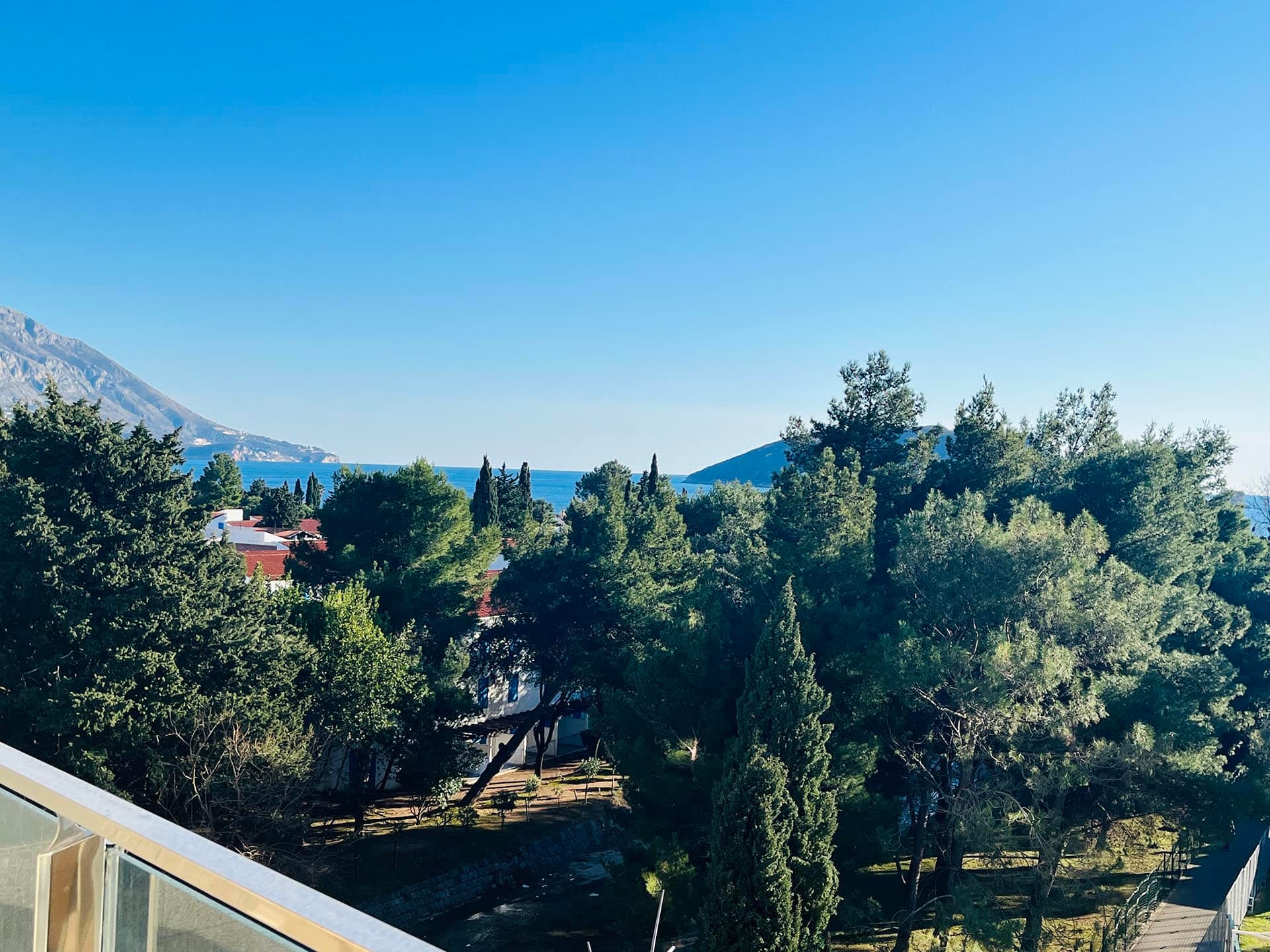 Karadağ, Budva’daki dairenin balkonundan görünüm: güneşli bir দিনে Adriyatik Denizi, yeşil park ve dağların panoraması.
