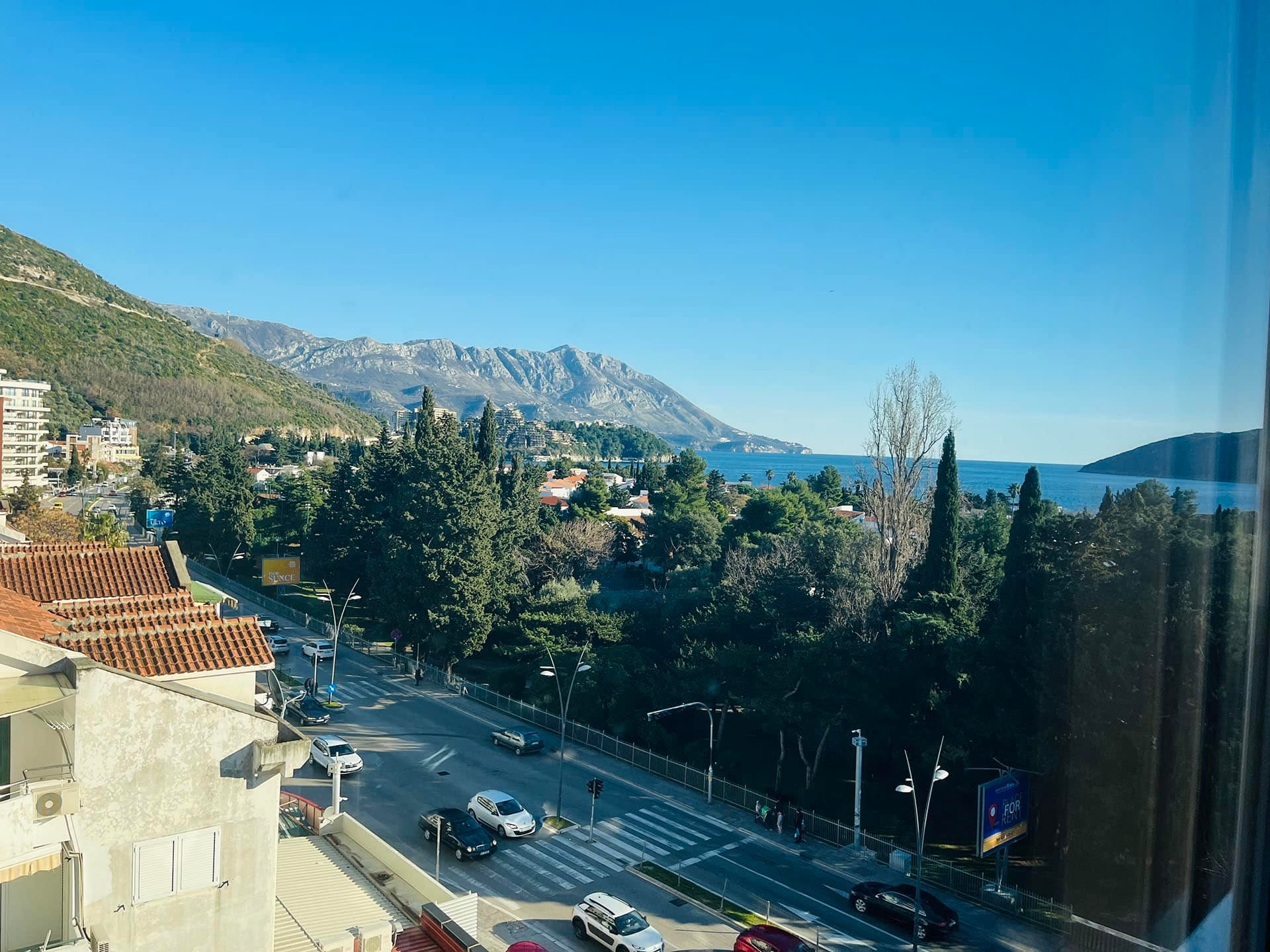 Budva, Karadağ’daki daire balkonundan görünüm: güneşli bir দিনে deniz, dağlar, yeşillik ve şehir sokağı panoraması.
