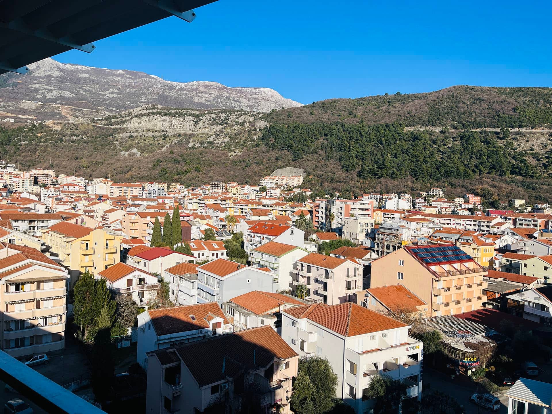 Karadağ, Budva’daki dairenin balkonundan panoramik manzara: kiremit çatılı şehir yapıları ve dağ manzarası.