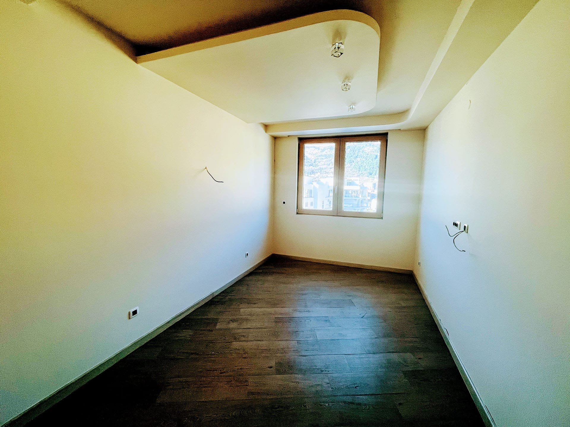 Karadağ, Budva’daki yeni dairede aydınlık oturma odası: tasarım tavan, koyu laminat ve pencereden manzara.