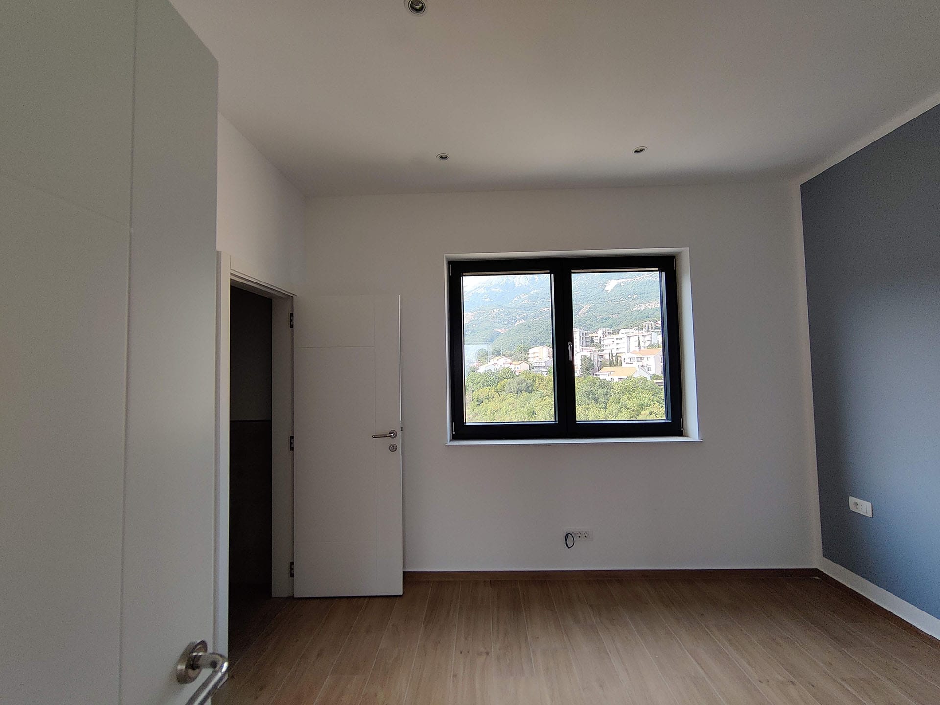 Svetla spavaća soba u stanu u Bečićima, Crna Gora: laminat, beli zidovi, akcentni plavi zid i pogled na planine kroz prozor.