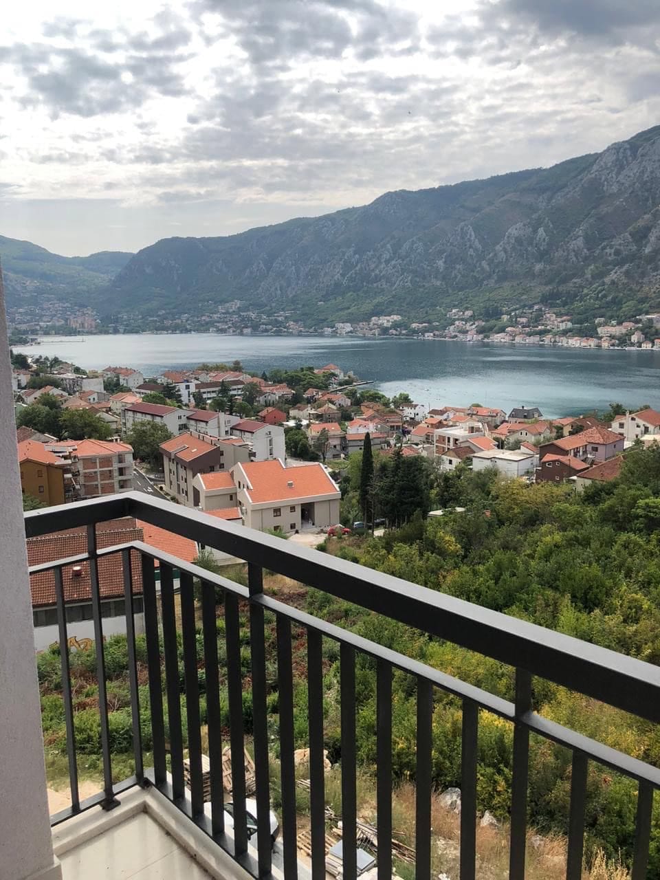 Pogled sa balkona stana u Dobroti, Crna Gora: panorama Bokokotorskog zaliva, planine i krovovi kuća od crepova.