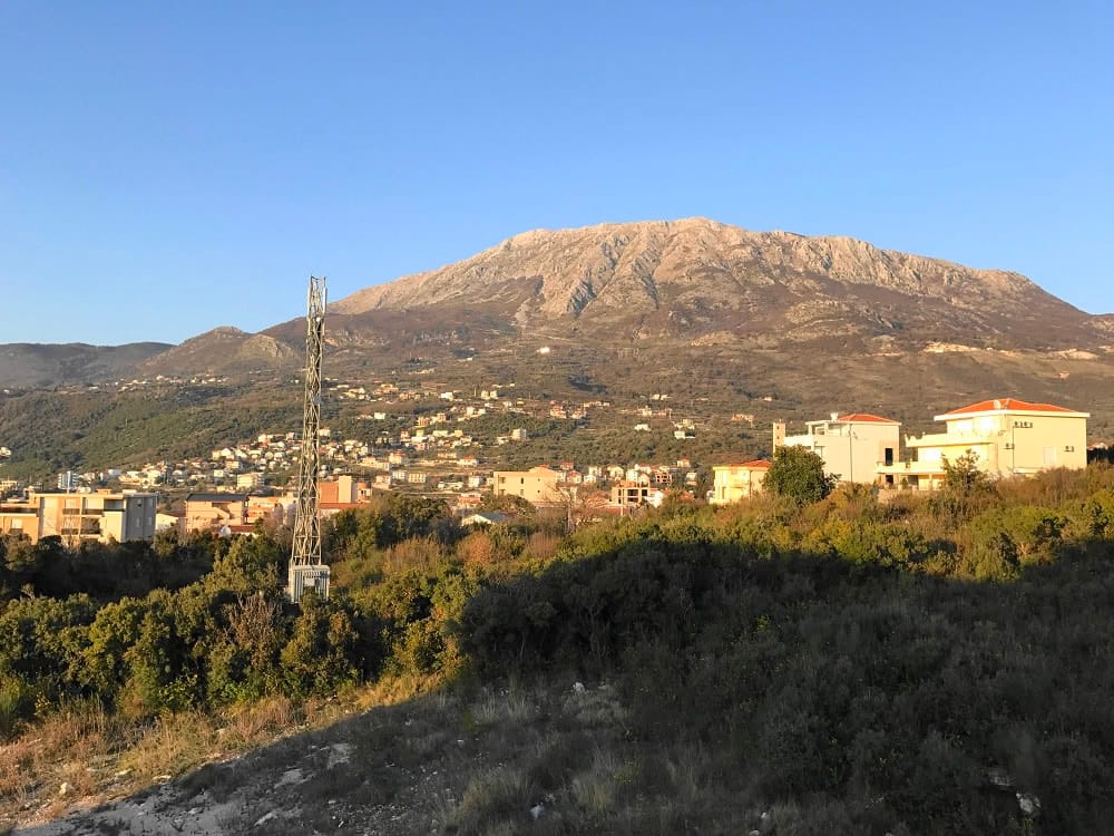 Panoramic view of the mountains and residential area in Dobra Voda, Montenegro: private houses, greenery, and a clear sky at sunset.