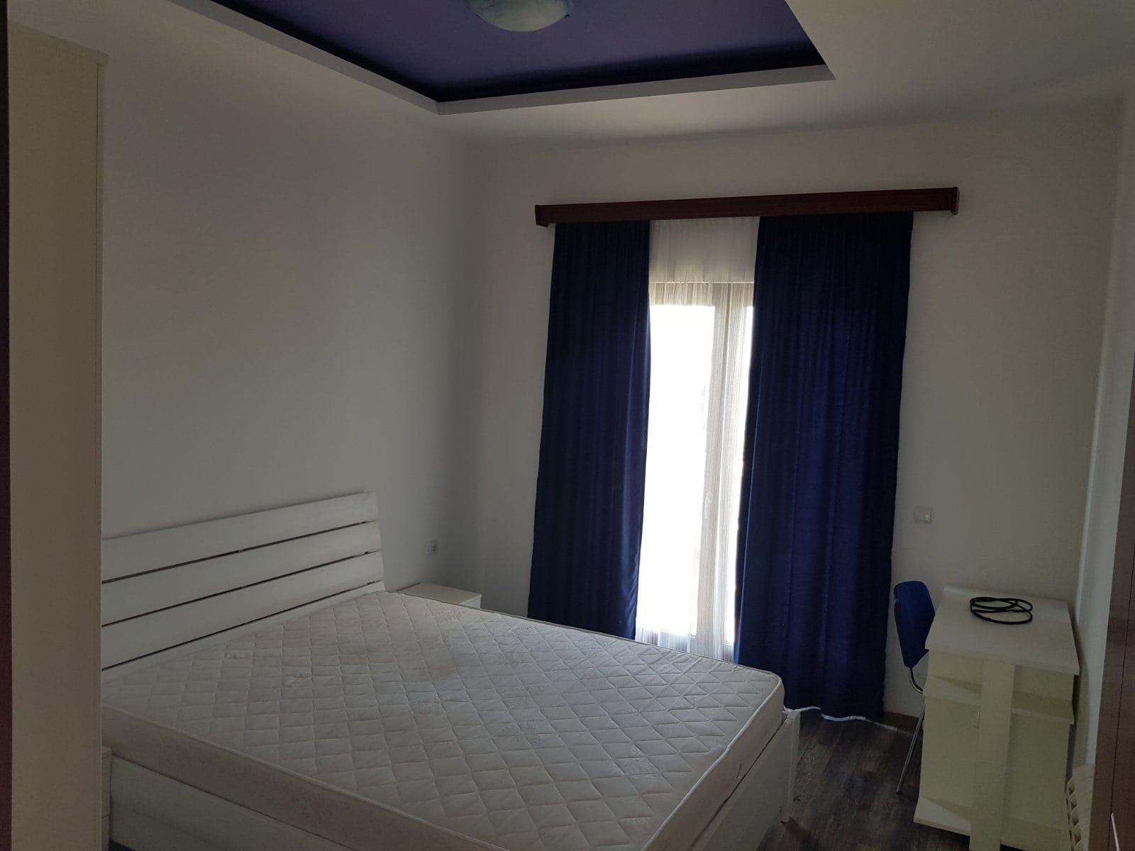 Svetla spavaća soba sa belim krevetom i plavim zavesama u kući u gradu Bar, Crna Gora. Moderan enterijer sa osvetljenjem.