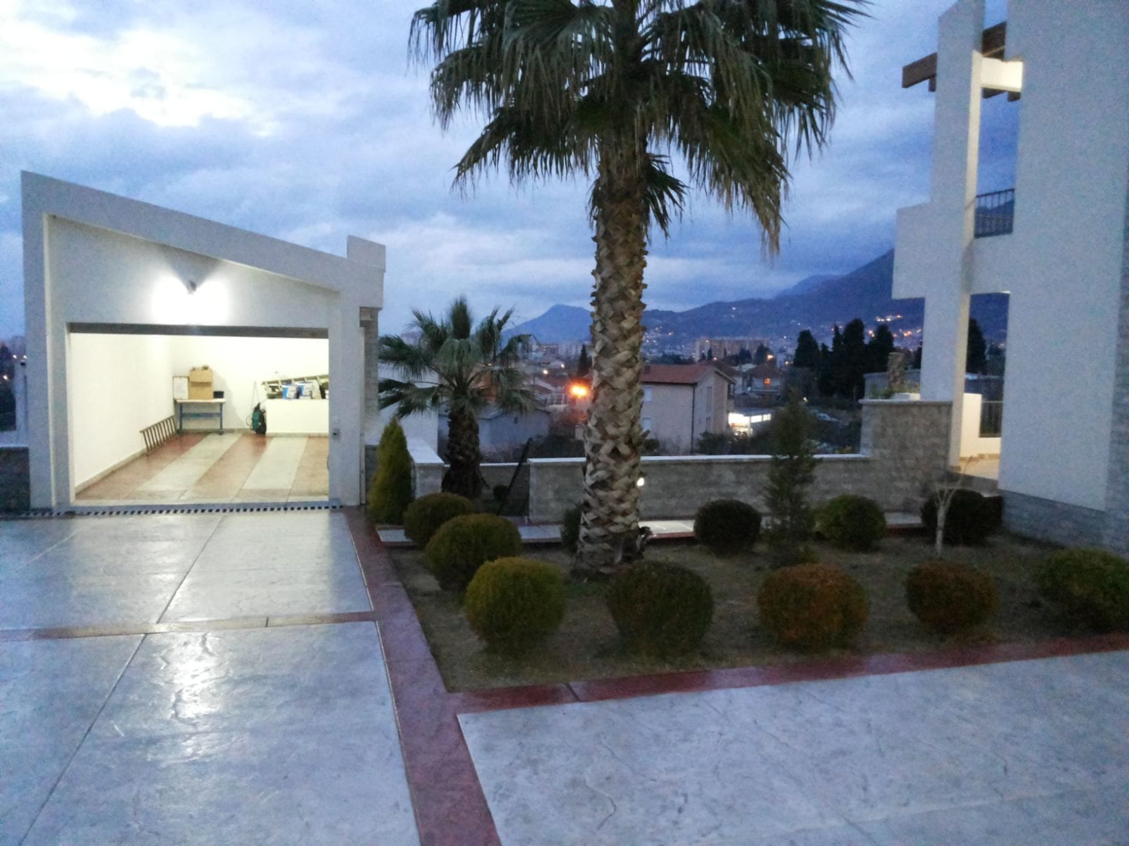Savremena kuća u Baru, Crna Gora: pogled na garažu, uređenu baštu sa palmom i panoramski pogled na planine uveče.