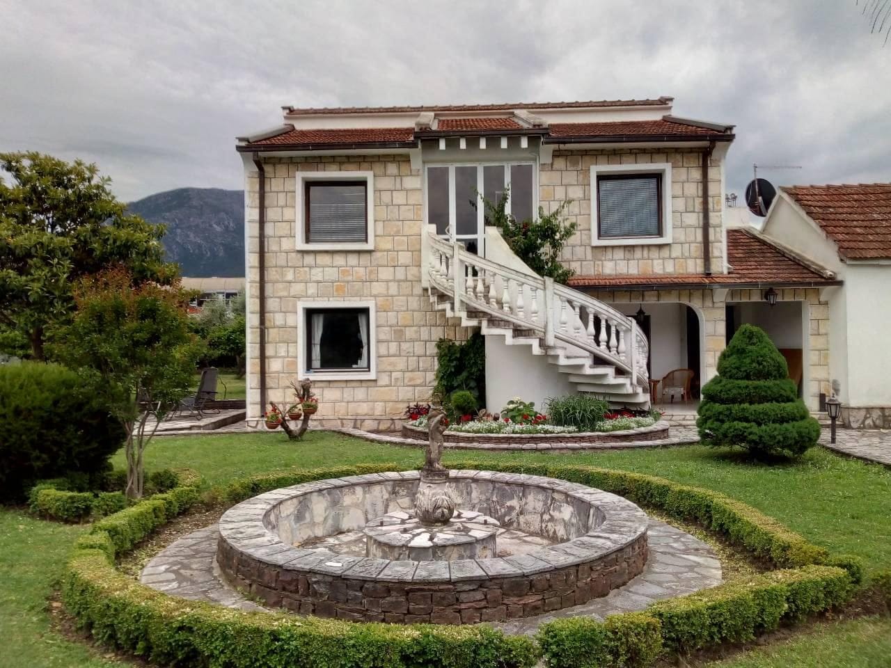 Dvospratna kamena kuća u Baru, Crna Gora, sa spoljnim spiralnim stepeništem, baštom i okruglom fontanom na placu.