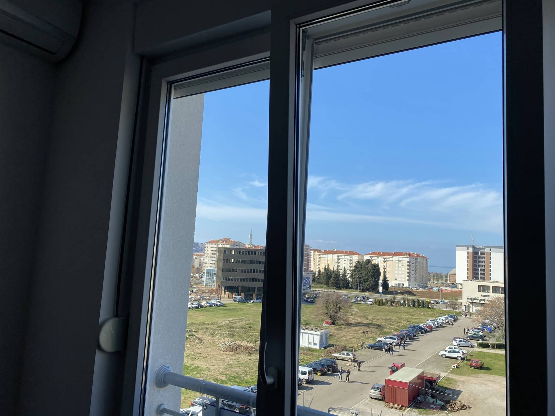 Pogled iz prozora stana u gradu Bar, Crna Gora: gradska izgradnja, parking i vedro nebo kroz plastični prozor.