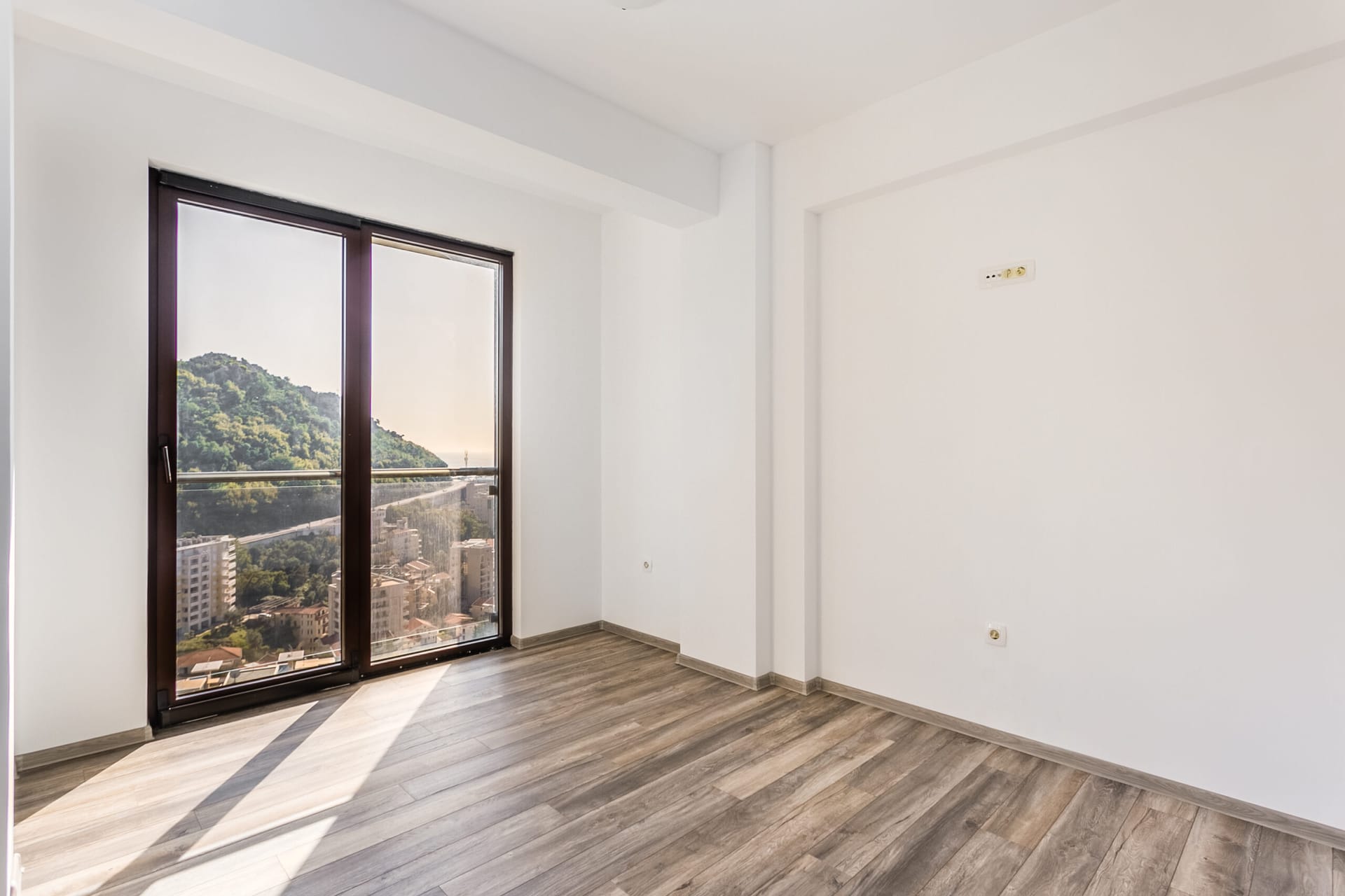Svetao dnevni boravak u penthausu u Rafailovićima, Crna Gora: panoramski prozor s pogledom na planine, laminat i beli zidovi.