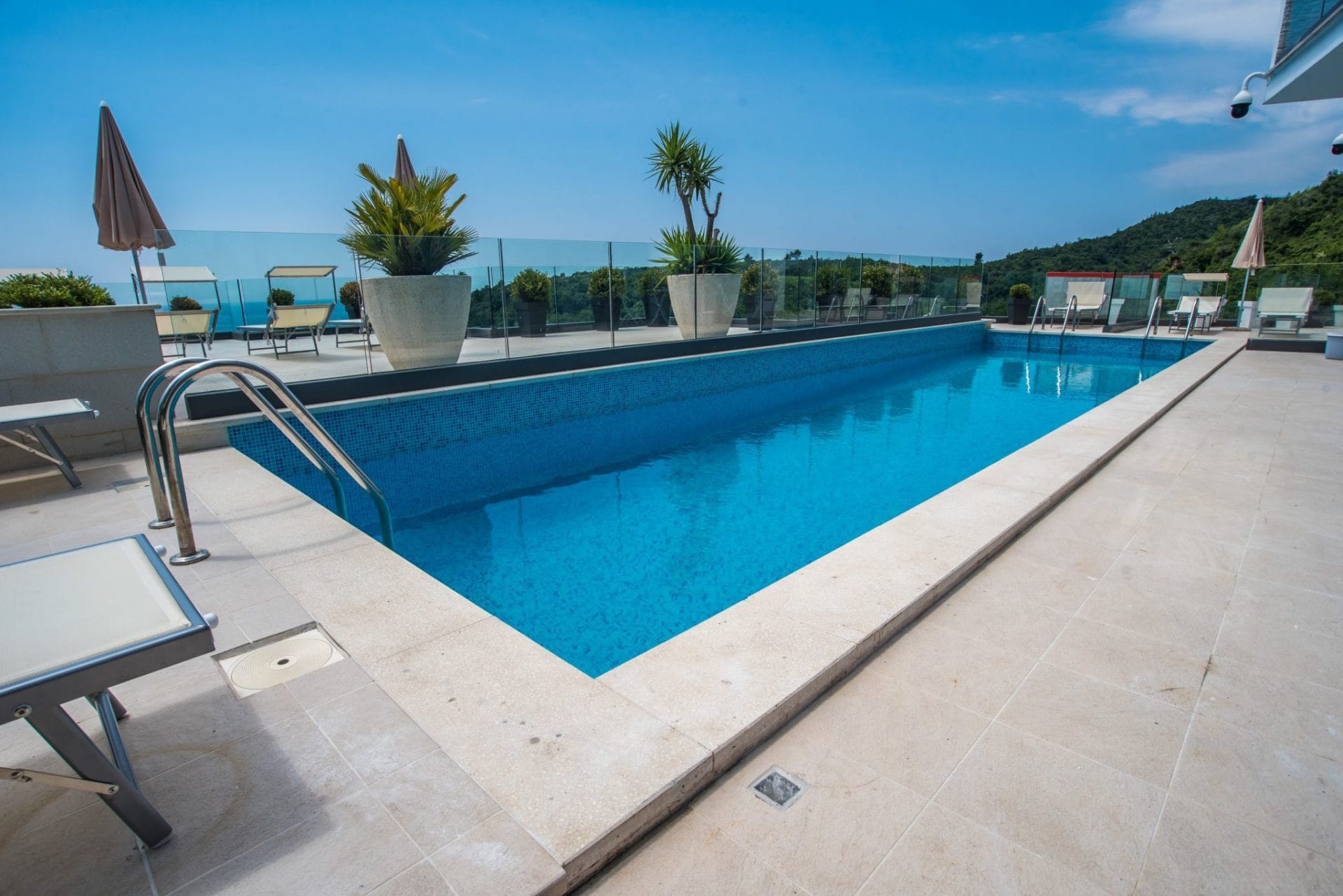 Open-air pool on the villa terrace in Budva, Montenegro, with sun loungers, glass railing, and mountain views.